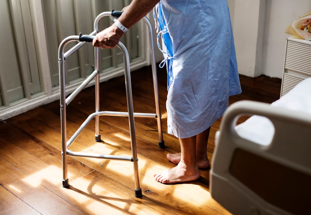 persoana varstnica in costum de spital care merge sprijinita pe un cadru de mers
