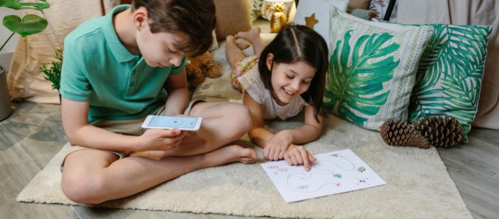 doi copii care stau pe podea, baietelul se uita pe telefon pentru a gasi un indiciu iar fetita se uita pe o harta