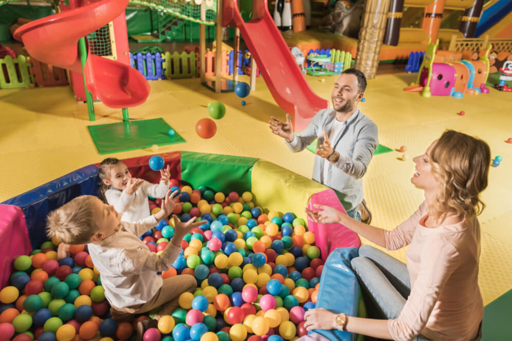 loc de joaca cu familia unde doi copii, fetita si baietel se joaca in piscina cu bile si mama si tata ii incurajeaza de langa si se joaca si ei