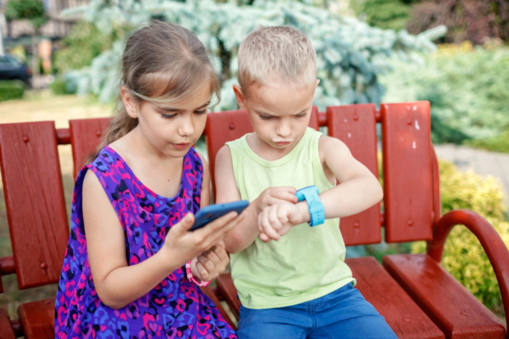 imagine copii care stau pe o banca unul are un smartphone iar celalalt un smartwatch la mana