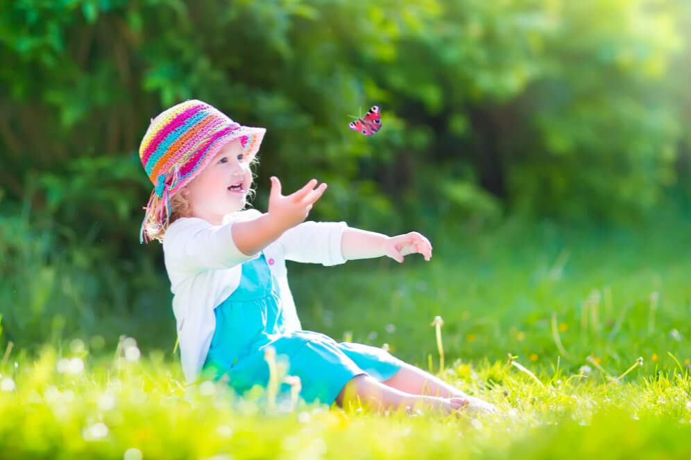 copil mic cu palarie multicolora pe cap, care sta in sezut pe iarba si se joaca cu un fluture