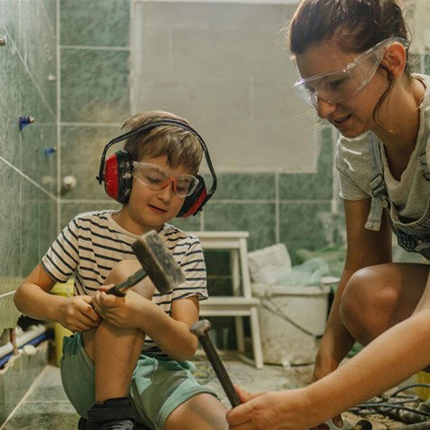 copil cu ciocan in mana si casti antifonice pe cap isi ajuta mama sa bata cu dalta in gresie pentru a o scoate