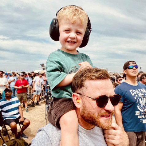 copil blond cu casti antifonice negre pe cap care sta in spatele tatalui la un eveniment in parc