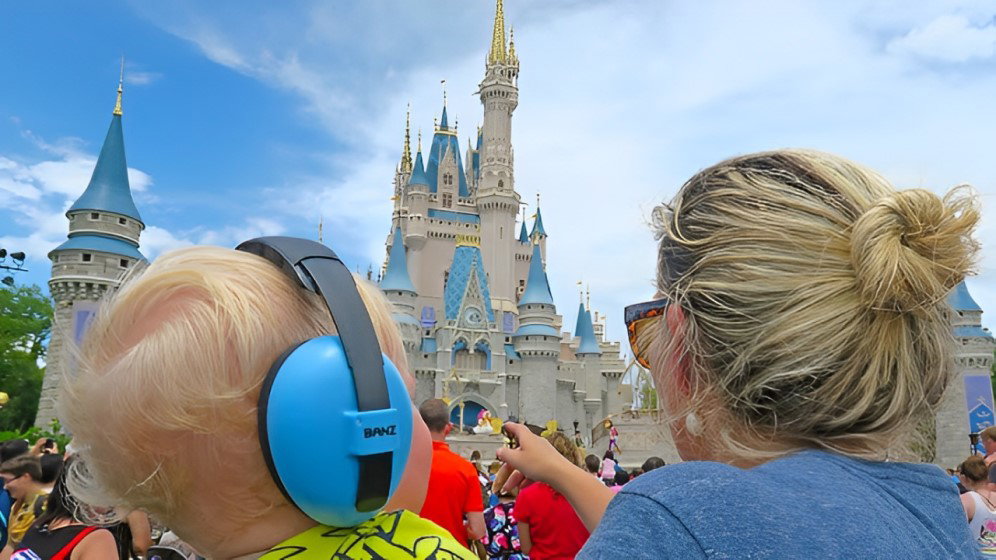 copil cu casti antifonice albastre marca Banz, in bratele mamei si se uita amandoi la un castel Disneyland