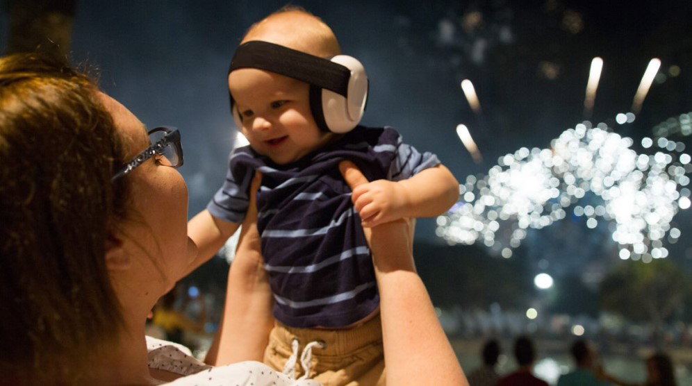 bebe cu casti antifonice in bratele mamei cu foc de artificii in spate